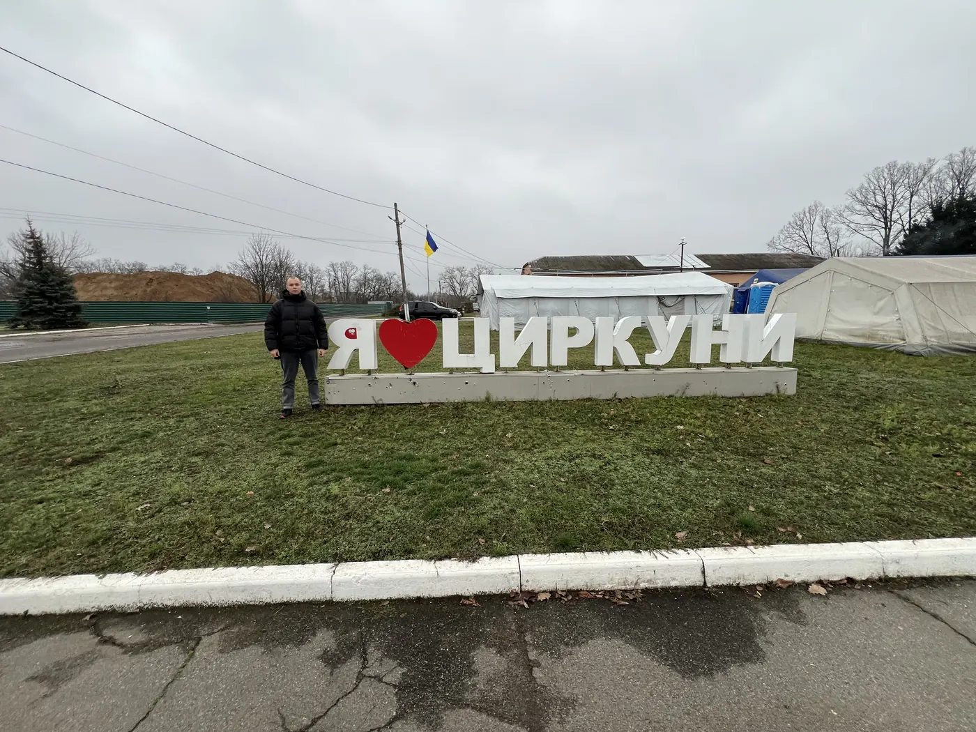 Чоловік стоїть біля великого напису «Я ♥ ЦИРКУНИ» край дороги; позаду — намети пункту незламності та український прапор під сірим зимовим небом