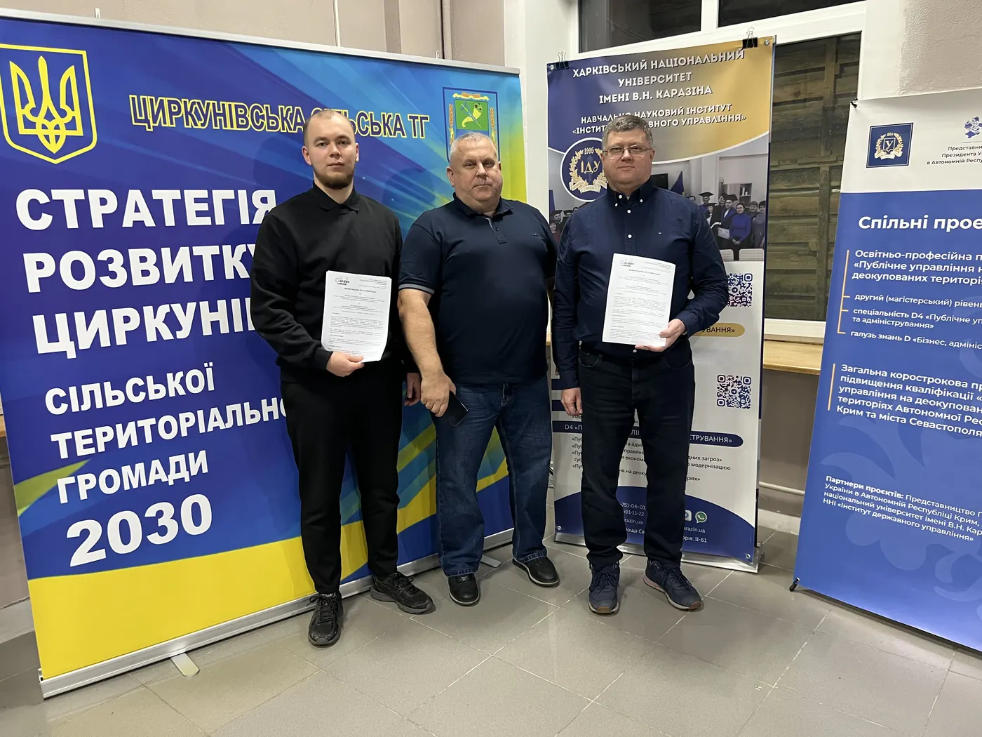 Троє учасників зустрічі стоять із підписаними документами в руках перед ролапами Циркунівської громади та Каразінського університету