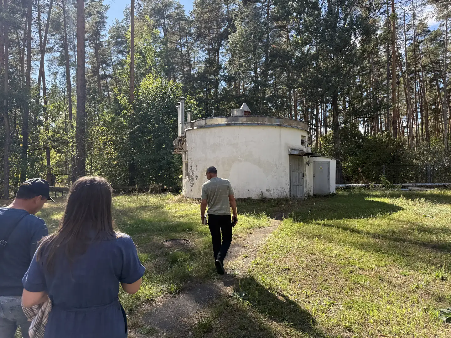 Люди йдуть стежкою серед сосен до круглої білої будівлі на території центру