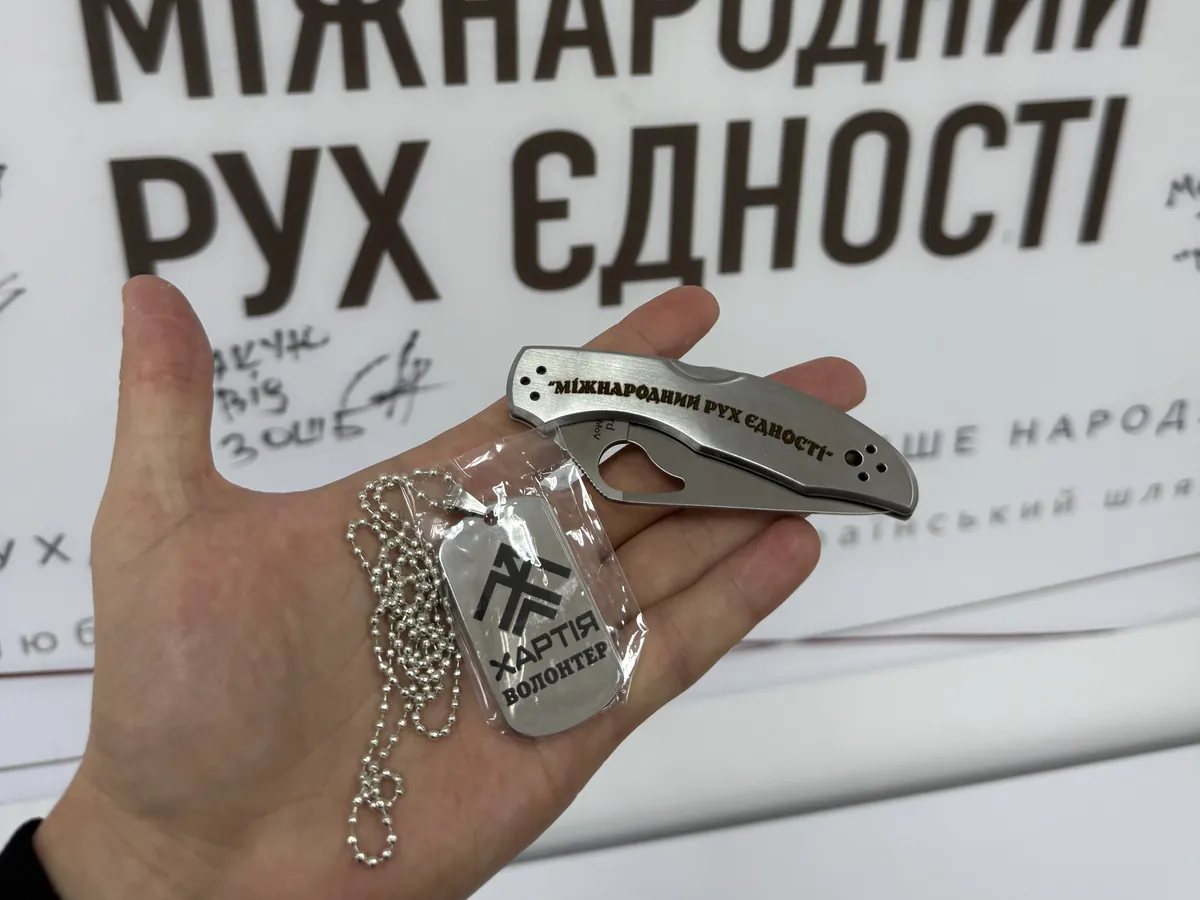 Рука тримає складаний ніж із гравіюванням «Міжнародний Рух Єдності» і жетон «Хартія · Волонтер» на тлі банера фонду з підписами військових