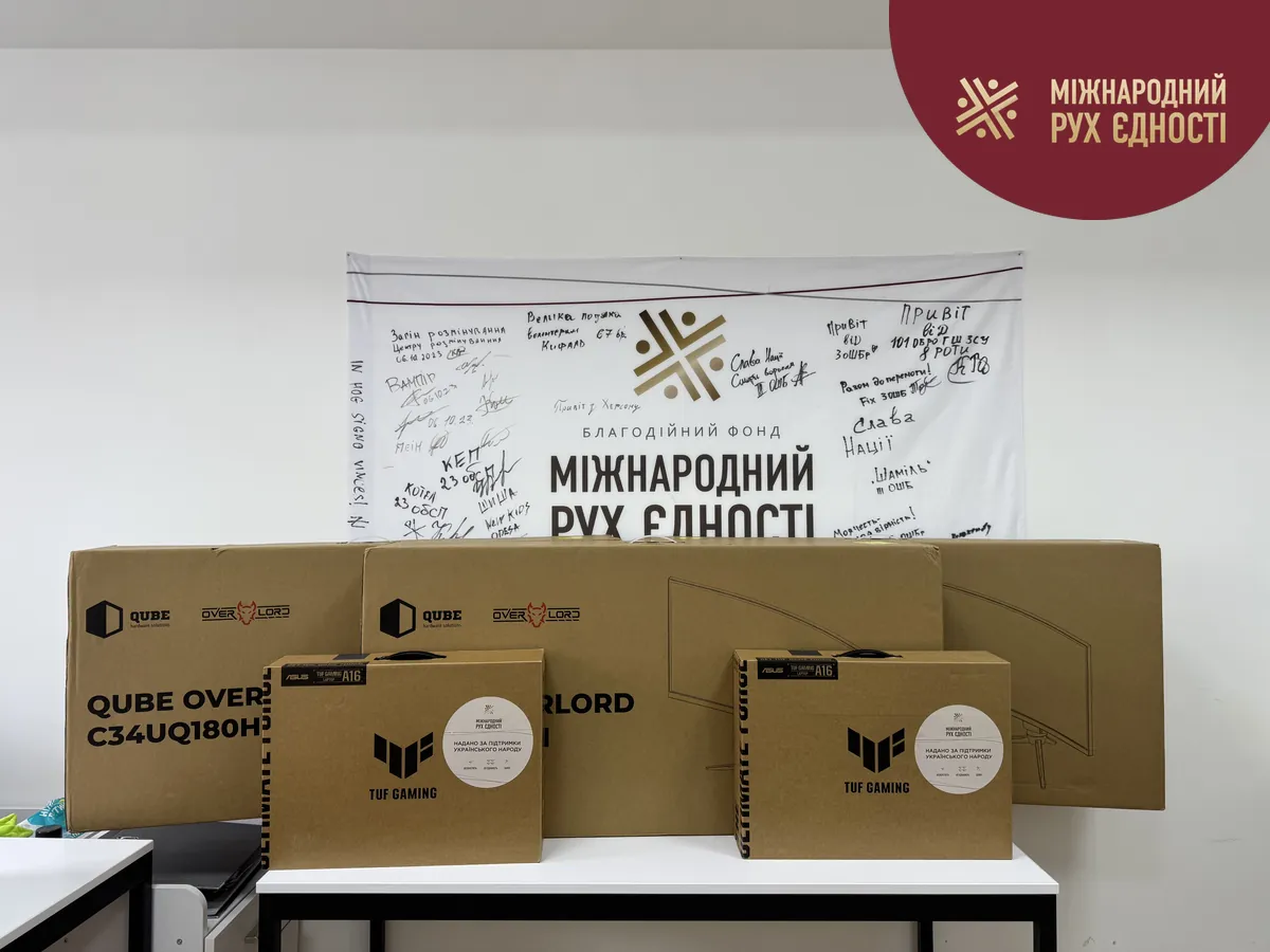 Коробки двох ноутбуків Asus TUF Gaming A16 і трьох моніторів QUBE Overlord на тлі банера благодійного фонду «Міжнародний Рух Єдності» з підписами підрозділів