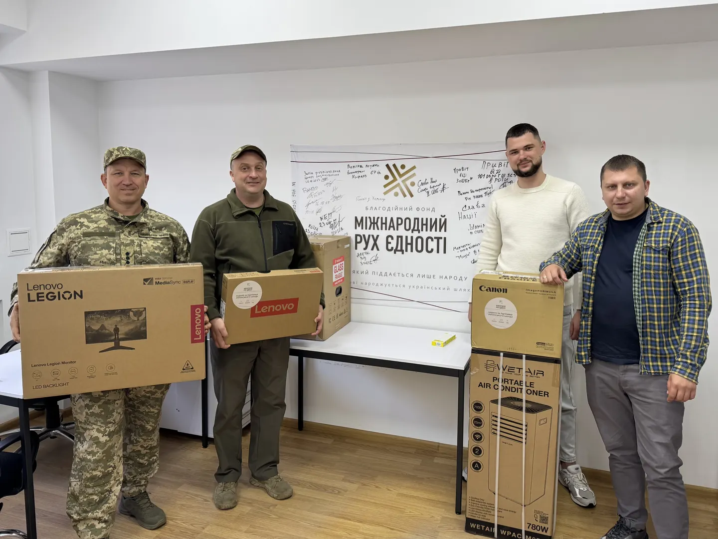 Двоє військових 101-ї бригади охорони Генштабу з коробками монітора Lenovo Legion і ноутбука Lenovo, поруч двоє цивільних біля коробок сканера Canon і кондиціонера WetAir, офіс фонду