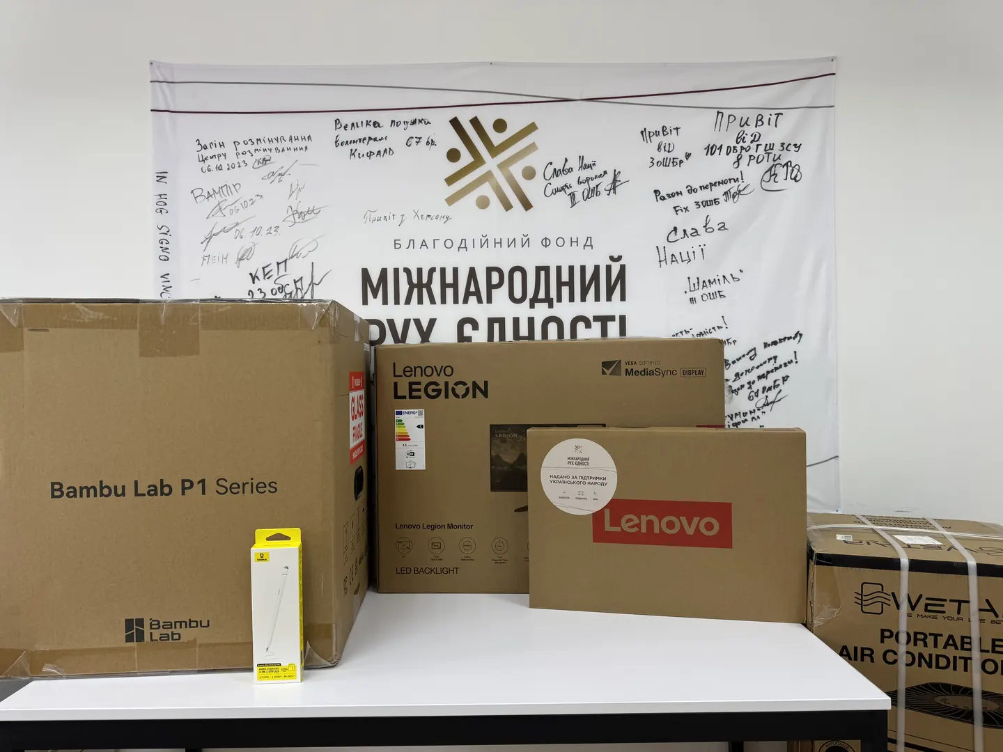 3D-принтер Bambu Lab, монітор і ноутбук Lenovo, кондиціонер WetAir і стилус Baseus на тлі банера благодійного фонду «Міжнародний Рух Єдності» з підписами підрозділів