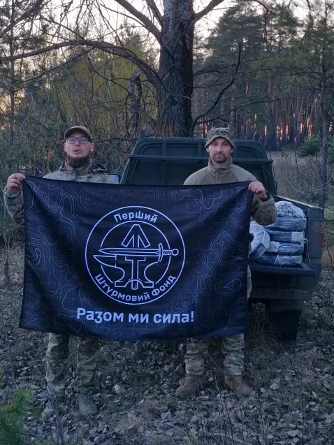 Двоє військових тримають прапор із написом «Перший Штурмовий Фонд. Разом ми сила!» на тлі пікапа в лісі, у кузові - бухти кабелю в плівці. Кадр із відеоподяки