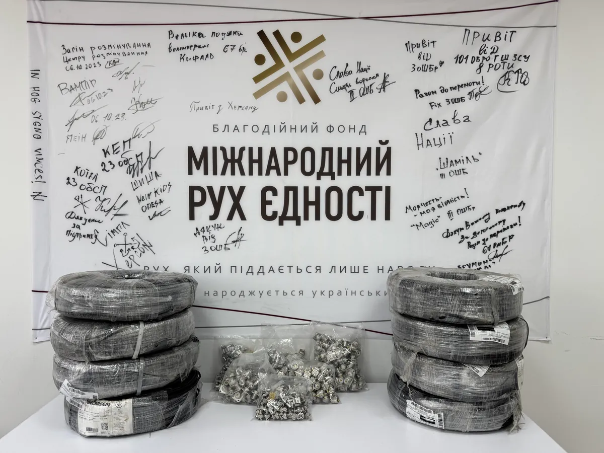 Вісім бухт коаксіального кабелю RG-58 у плівці й пакети з роз’ємами на столі на тлі банера фонду «Міжнародний Рух Єдності» з підписами підрозділів
